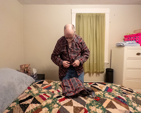 Color photo of a man in a bedroom buttoning up his pajama top.
