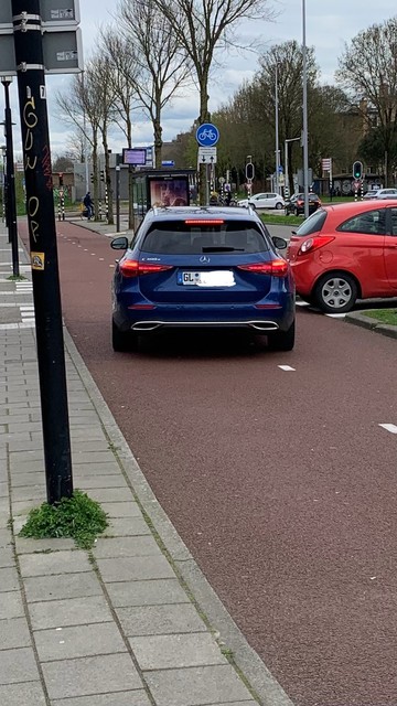 A blue Mercedes car with German licence plate on a bike lane next to a sidewalk, with traffic and pedestrian infrastructure visible in the background.