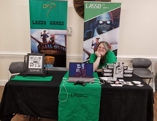 The lasso games booth set up for herdcon! Our banners are in the background that feature our logo and Minilaw and Levantera (Highway rampage is on there too but you can't see it from the picture). Someone sits behind the table smiling for the camera. On the left we have a QR code for our socials, in the middle a laptop playing Highway Rampage and finally on the right is Random Quest set up as if to be played. This is on a black table with a green runner down the center with our Logo embroidered on it. 