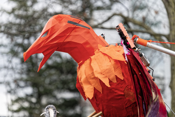 Bright orange mechanical bird being held up from behind in front of defocused trees.