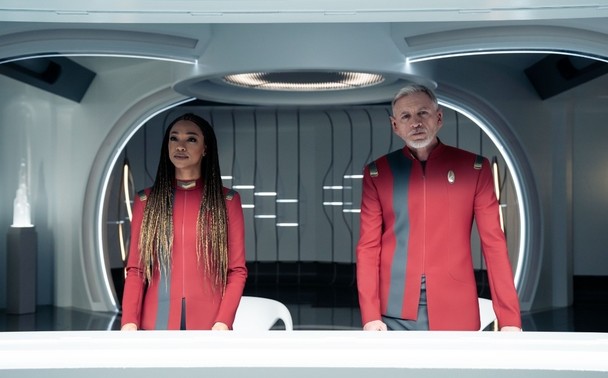 Capt Burnham (Sonequa Martin-Green) and Captain Rayner (Callum Keith Rennie) stare out towards the camera. Behind them is a rounded room in sleek shiny futuristic Star Trek fashion.