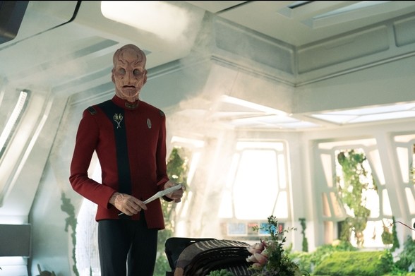 Alien First Officer Saru (Doug Jones) stands in a room full of green plants, while holding a info pad.