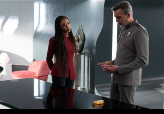 Capt Burnham (Sonequa Martin-Green) states with her head tilted, in the direction of Admiral Vance (Oded Fehr), as he looks down at something he is holding in his hands.