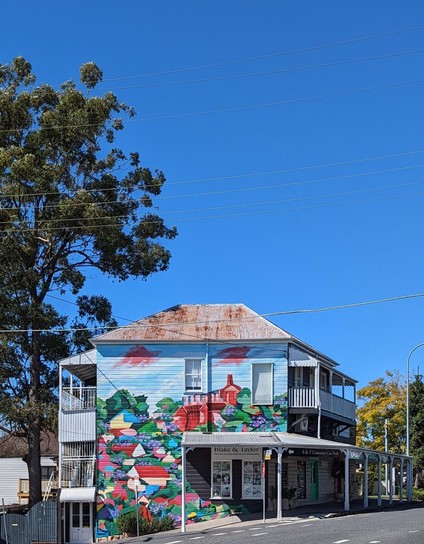 A "Queenslander" with rusty tin roof, and a leafy village depicted across the whole side of the building.  Trees, blue sky above.