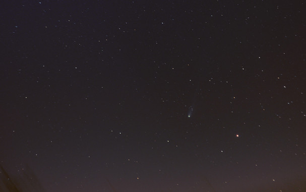 Night sky, with colorful stars and ugly noise between them, fading into brownish muck near the horizon, with some dark streaky artifacts of foreground objects blurred by stacking images aligned on the stars. The comet is toward the lower right, just above the worst of the muck with a green central coma and slightly bluegreenish tail fading to toward the upper right