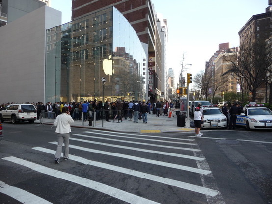 glass shop building with Apple logo: around the building a line of people. In the foreground: a zebra crossing. On the right in the picture: NYPD police cars.