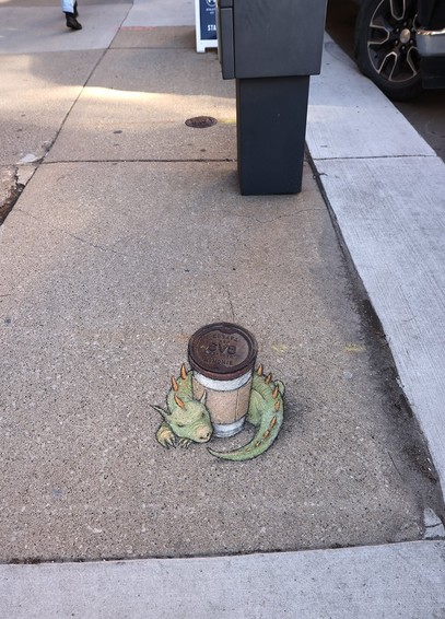Street Art. Zeichnung auf dem Boden.
Ein kleiner grüner Drachen mit gelben Stacheln auf dem Rücken, umringt einen Kaffeebecher mit Deckel.