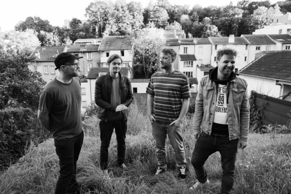 Schwarz-weiße Fotografie der vierköpfigen Band "Paper Planes" aus Offenbach. Sie stehen im Gras. Im Hintergrund von großen Bäumen eingefasste Häuser.