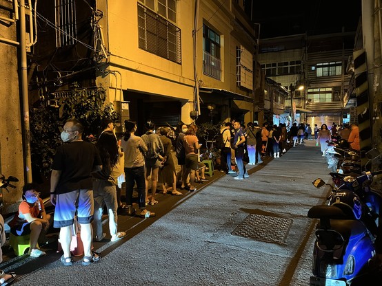 A narrow street at midnight in Taiwan with residential buildings on either side.  On the right scooters are parked all the way down, while on the left is a long queue of people both standing and sitting on their own small chairs, waiting to buy spring roll skins.  The street is lit by a combination of orange street lighting and the cool direct light of a scooter.