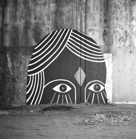 A black and white film photo. A mural under a downtown Austin bridge of a person with only half their face visible protruding from the ground. Tuesday, April 2, 2024.