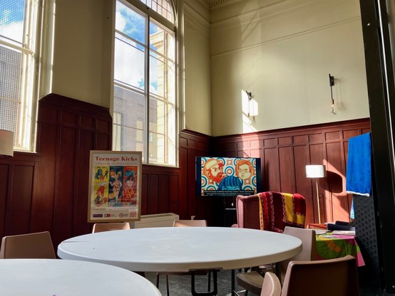 Image of a high-ceilinged wood-paneled sunlit room with tables and armchairs, with a TV screen and sign depicting the Manchester Uni illustrated project “Teenage Kicks: Girls Growing Up in Britain 1956-1974”