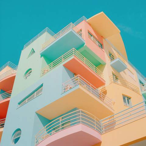 Upwards angle on a photo of a high-rise apartment building, with many balconies. The colours are very weird, like colours you might wear to the beach. 
