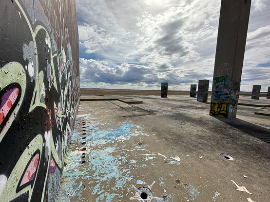 Graffiti on a wall has spilled blue and white swirls on the concrete base, mirroring the blue sky with white clouds streaming over the site