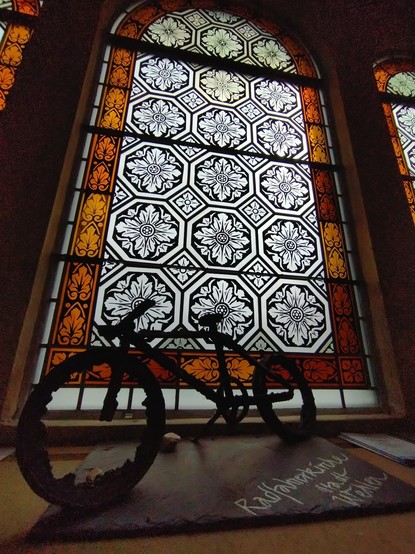 Kirchenfenster mit Bleiverglasung, orangefarbener Rand mit Blattmusterung, innen weis-schwarze Achtecke mit Blütenmuster. Im Vordergrund steht auf dem Fenstersims eine Fahrradskulptur aus rostigem Eisen auf einer Platte, die die silberne Beschriftung trägt: Radfahrerkirche Stadt Wehlen. Daneben liegen ein paar Flyer.