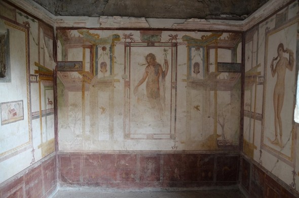 Fresco of Dionysos, photographed from the entry to the room. The god is standing, holding his thyrsos in one hand and probably a cup in the other but it has sadly been damaged. He is depicted with long, curly hair flowing over his shoulders and the same complexion as Venus, underlining his effeminacy.