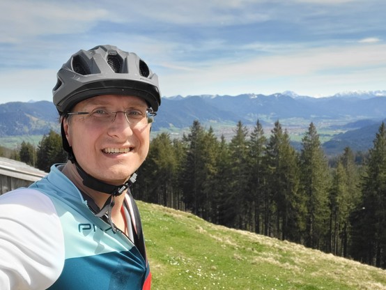 The image captures a moment of outdoor serenity and adventure, featuring a man who appears to be in his mid-forties, exuding a sense of joy and contentment. He is dressed in attire suitable for outdoor activities, most notably wearing a bicycle helmet, which suggests a penchant for cycling or similar recreational pursuits. The helmet, coupled with glasses, underscores a commitment to personal safety and protection. His face is adorned with a smile, signaling enjoyment or satisfaction with his c…