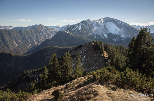 The mountains, standing majestic with their summits reaching towards the sky, are adorned with a delicate dusting of snow that highlights their rugged contours. The scene is richly populated with diverse vegetation, including dense clusters of trees that appear to be predominantly conifers, suggesting the photograph might have been taken in a region similar to the alps or a highland area known for its temperate coniferous forests. 
