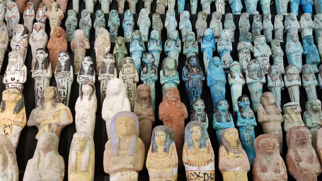 Approx 7 rows of Ancient Egyptian Shabti statues. Each row has over ten. Some are glazed, others not. Some are coloured turquoise, or brown, and some have panted detail like coloured hair, outlined eyes, and Hieroglyphs. 

