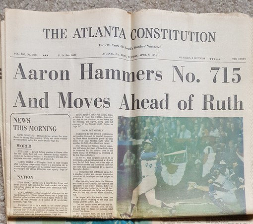 The front page of The Atlanta Constitution newspaper dated April 9th 1974. Headline reads "Aaron Hammers No. 715 And Moves Ahead of Ruth," and the photo is a shot of Henry 'Hammering Hank' Aaron hitting that home run.
