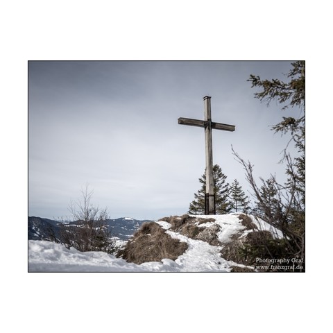 A serene winter scene captured in this photograph showcases a wooden cross standing on a snowy hill. The landscape features a backdrop of snow-covered trees and a mountain range in the distance. The dominant colors of white and grey create a peaceful and cold atmosphere. Two trees are visible in the foreground, one on the left side and the other on the right side of the image. The image also includes a wooden pole with a sign attached to it. Overall, this image conveys a sense of tranquility an…