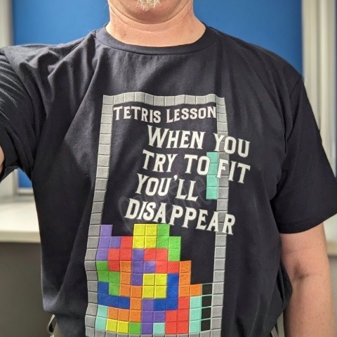 Eine Person steht vor einer blauen Wand und trägt ein schwarzes T-Shirt mit einem bunten Tetris-Spiel-Design und dem Text ‘Tetris Lesson: When you try to fit, you’ll disappear’.