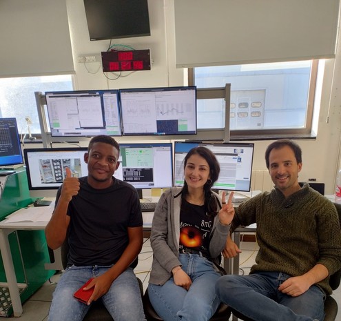 Three people sitting in front of computer monitors with graphs and data, smiling and gesturing thumbs-up and peace sign, in an office-like setting with a digital clock displaying the time on the wall above. Control room of IRAM 30m telescope.
