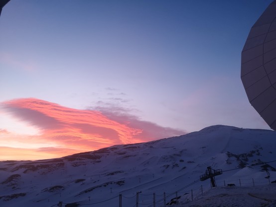 Snow-covered mountain slopes with ski lifts at dusk, under a sky with pink clouds. A part of a large, curved architectural structure is visible on the right, which is part of the 30m telescope. © Salvador Sanchez 