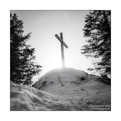 A serene and tranquil scene captured on a snowy hill, featuring a wooden cross standing tall against the white backdrop. The landscape is dominated by shades of white and grey, with a hint of a soft grey sky in the background. Two trees frame the cross, one on each side, their bare branches reaching towards the sky. The image is in black and white, adding to the overall sense of peace and stillness. This breathtaking winter scene evokes a sense of spirituality and connection with nature, making…