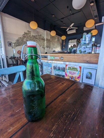 Cafe scene. 
Grolsch bottle on table foreground. 
Posters advertise events at the counter. 
Yellow globe lights hang from the black ceiling. The white wall is decorated with a large abstract black line pattern.