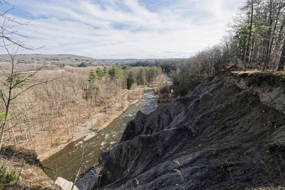Wide angle view of a creek with white rapids,  several ridges can be seen going down from the cliff I am on.  On the other side there are mostly deciduous trees that haven't leafed out yet,  merging into a background with gentle rolling hills on the horizon and a mostly cloudy sky.