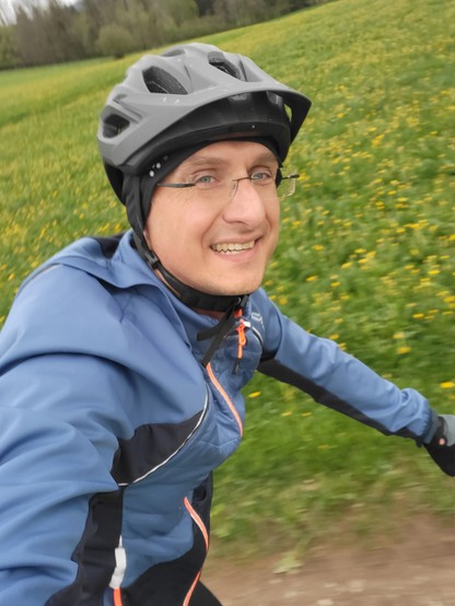 A middle-aged man with a smile on his face is shown riding a bicycle in an outdoor grassy area. He is wearing a black bicycle helmet for personal protective equipment. The man is wearing a blue jacket and appears to be enjoying his ride, as indicated by his smile. The dominant colors in the image are black and yellow, with the accent color being a shade of green. The image also includes objects such as footwear and a Bicycle helmet. The man's face is visible, showing a male with an approximate …