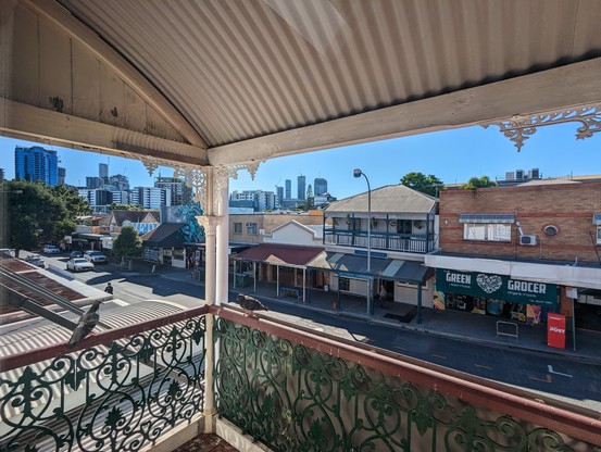Looking out from a verandah with a tin roof and scrollwork railings. 
In the foreground, Queenslanders and low buildings. In the distance, the CBD skyline  blue sky 