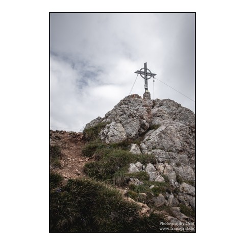 A serene and spiritual scene captured in this image showcases a white cross placed on a large rock in the outdoors. The rock is surrounded by natural elements such as grass and dirt, with a cloudy sky in the background. The composition is peaceful and harmonious, with the cross symbolizing faith and hope. The dominant colors in the image are white and grey, creating a tranquil atmosphere. The landscape features mountains in the distance, adding to the picturesque setting. Overall, this image co…