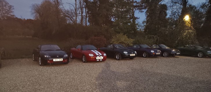 A wide shot at dusk with several MG F and MG TF sports cars parked on a gravel car park with trees behind.