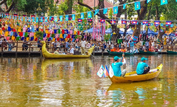 Photo shows golden coloured small boats on a mirror clear river in Siem Reab, Cambodia. One boat is filled with partying family ready for a trip on the river. An other have two crew staff keeping an eye out for security and cleaning tasks. On the river side there are crowds of celebrating people wearing their best, colourful decorations and Cambodian flags. In the background there are booths selling food, drinks snd celebration relating items. It represents a snapshot of the grand popular celebration and holidays being khmer New Year.