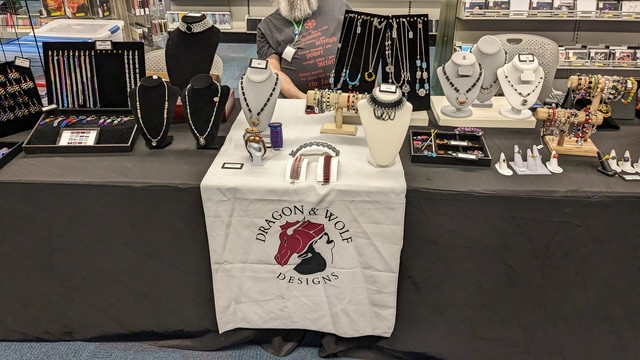 Vendor display of handmade jewelry. A white banner with the Dragon & Wolf Designs logo hangs in front of the tables. 