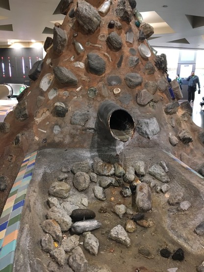 Sculpture of a conical pie of rocks and debris, with a pipe sticking out of it discharging a trickle of water