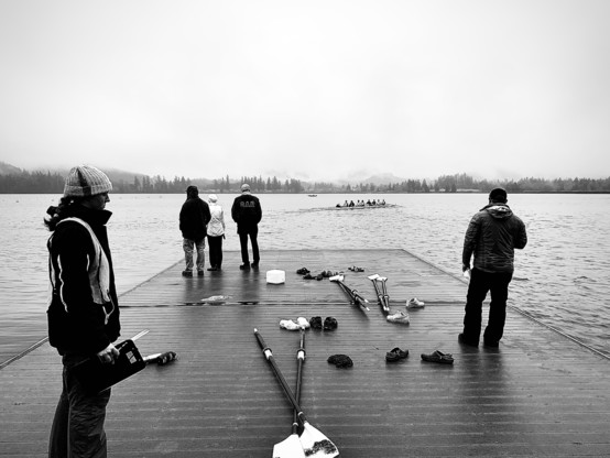 A black and white image of a dock at a lake with some people standing on it, and in the distance an eight person rowing skull leaving the dock. 