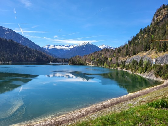 A serene lake is captured in the image, with majestic mountains towering in the background. The scene is set under a clear blue sky with fluffy white clouds adding a touch of tranquility to the landscape. Surrounding the lake are trees and a building, creating a picturesque view of nature's beauty. The mountains in the distance are part of a snow-capped range, enhancing the overall scenic beauty of the location. The image exudes a sense of peace and tranquility, making it a perfect representati…