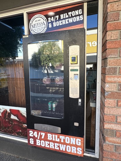 Photo of a vending machine, installed on the shop window of a specialty small goods and international groceries store, with the sign saying “24/7 Bolton and boerewors”. Inside the machine is sausage and soda.