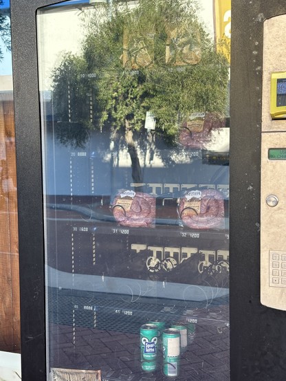 Close up photo of a vending machine, installed on the shop window of a specialty small goods and international groceries store, with the sign saying “24/7 Bolton and boerewors”, and inside  the somewhat empty machine - sausage and cans of soda can be clearly seen.