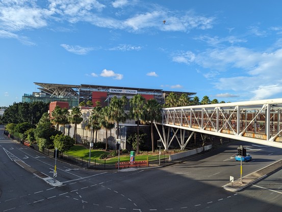 Bridge across road heading to stadium, which is large and Orange and surrounded by mature palm trees 