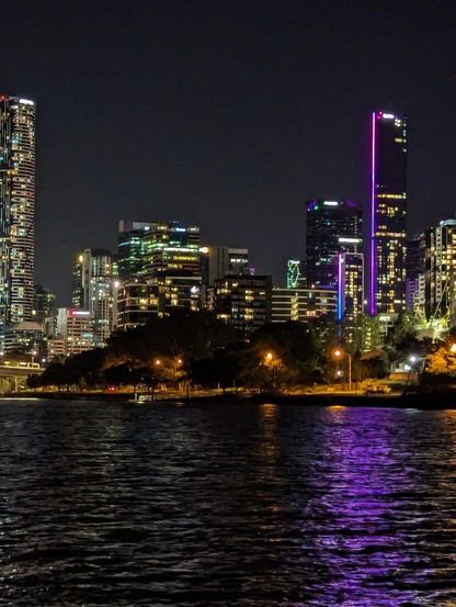 Cityscape across the Brisbane River showing a tall building with a vertical neon strip forming a purple reflection across the ripples of the river towards the viewer
