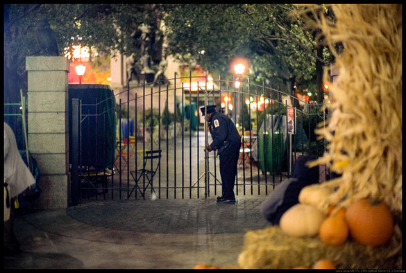 A policeman closes an iron gate and locks it.  The gate leads to a small park with chairs and tables.  Blurry pumpkins and straw are in the foreground.