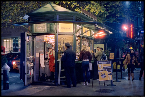 A small shop selling espresso at night.  Several customers stand in front while a worker stands framed in an open doorway.
