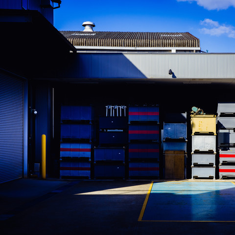 The photograph captures a corner of a factory where the sunlight casts sharp shadows across the scene. A stack of bright blue and red crates adds a pop of color in the otherwise dimly lit area. These containers are neatly arranged against the side of the factory, which is constructed of metal and concrete with a corrugated roof. The distinct lines of shadow and light play across the ground and the crates, highlighting their shapes and the orderly nature of the workspace.
