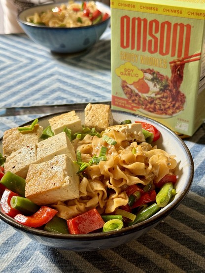 A bowl of cooked noodles with veggies and tofu. The dish looks very colorful, with bright red bell peppers and green peas. Near the dish is the empty box of Omsom noodles. 