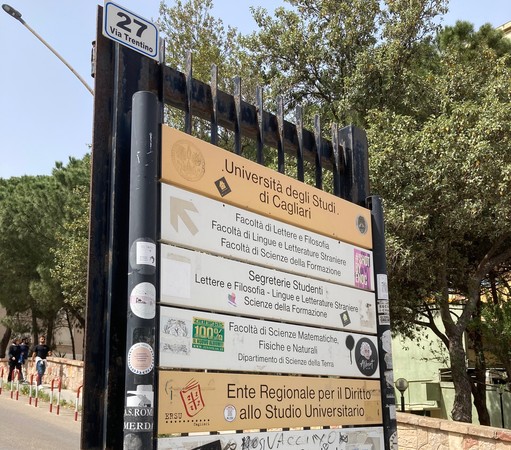 A sign at the entrance of the campus of the university of Cagliari. The sign is rather scruffy, with stickers and tags all over it, but you can make out the Facoltà di Lettere in the list. The landscape behind is mediterranean trees, in bright sunlight, already looking dry despite it being only early Spring.