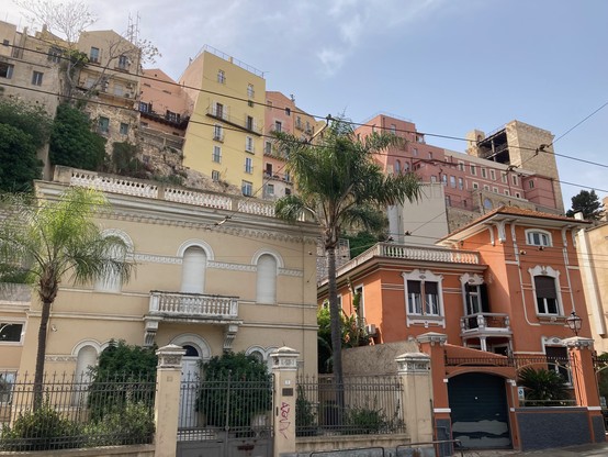 A view of the old town, taken from below. The foreground is a line of Italianate villas, looking rather fancy and freshly painted. The background, above, shows the jumbled houses of the old town along a cliff, near the castle. Pale pastel colours, in a sunny sky. 