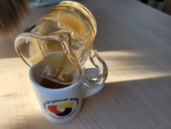 A white, 3 ounce, ceramic espresso (demitasse) cup with a shot of espresso, on a maple laminate desk surface illuminated by the morning sunlight. The blue text on the cup reads "Cyber-Physical Systems Research Center" and wraps in a circle around the logo, which looks like a loosely interlaced, rounded triquetra with thick knots in solid red, yellow, and blue (each its own color).

Resting on its side on the top of the cup is a 3-ounce Revolution Dual Spout Shot Glass, which was used to pour the espresso shot into the cup, and which still has some residual espresso crema on the inside.

A portion of a tail of a cat appears in the upper-left.